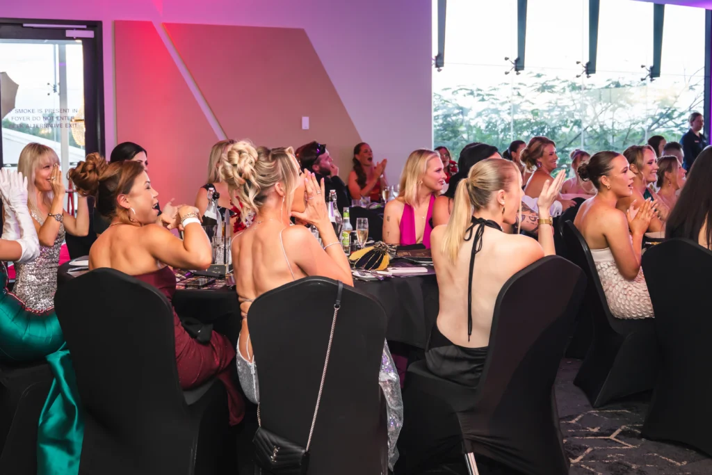 Guests in formal evening wear seated at tables applauding during the Magnify Her Gala
