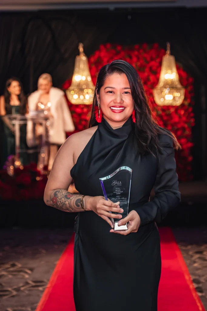 Woman in a black evening gown holding an award on a red carpet stage with a red rose backdrop at the Magnify Her Gala