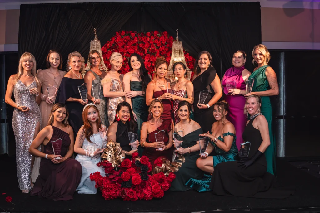 Group of women in formal evening gowns holding awards on stage at the Magnify Her Gala, posed in front of a red rose backdrop.
